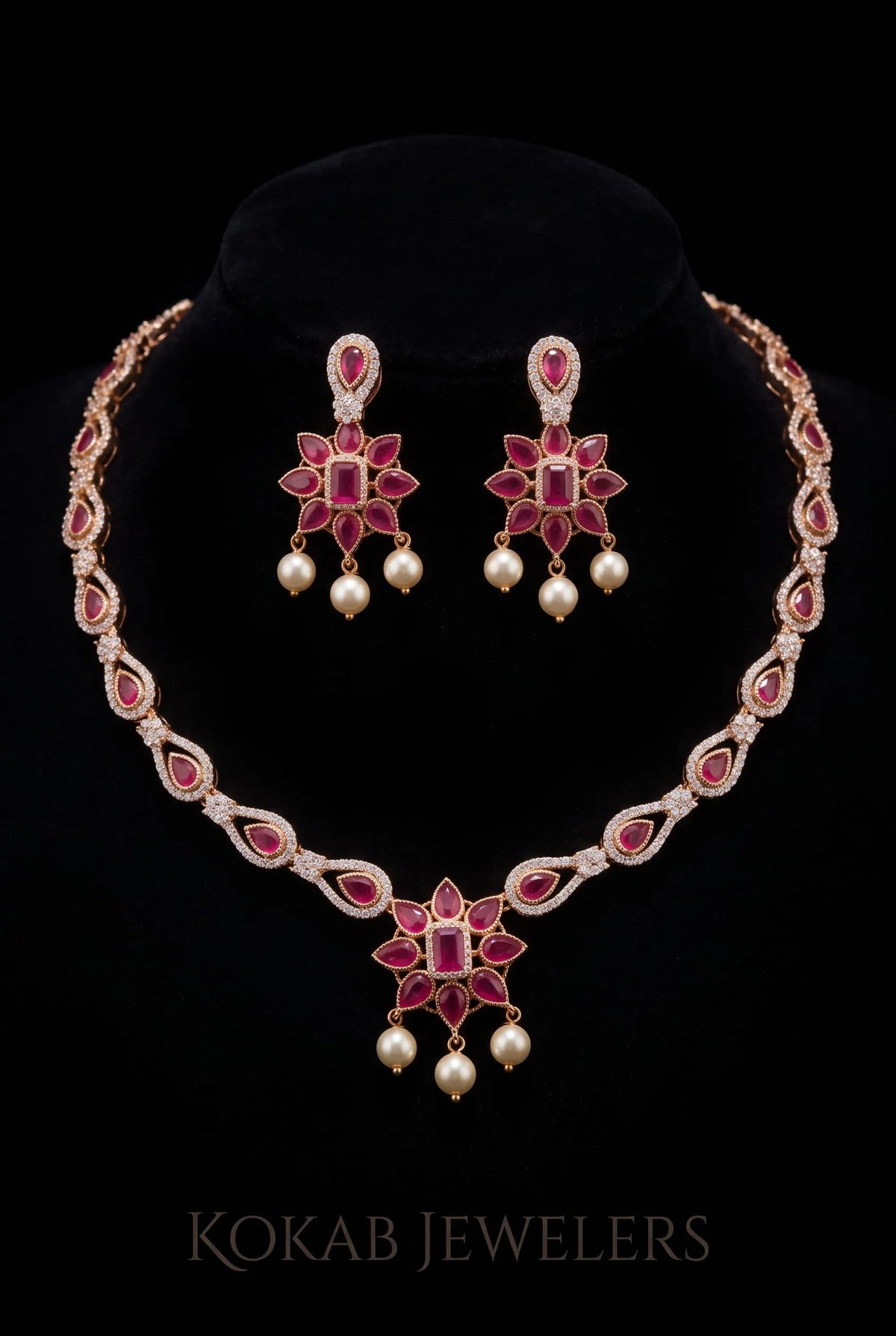 A traditional Indian jewelry set featuring a necklace and matching earrings with pink gemstones, white pearl-like drops, and gold-toned settings, displayed on a black velvet stand against a black background. The brand name 'KOKAB JEWELERS' is visible at the bottom.