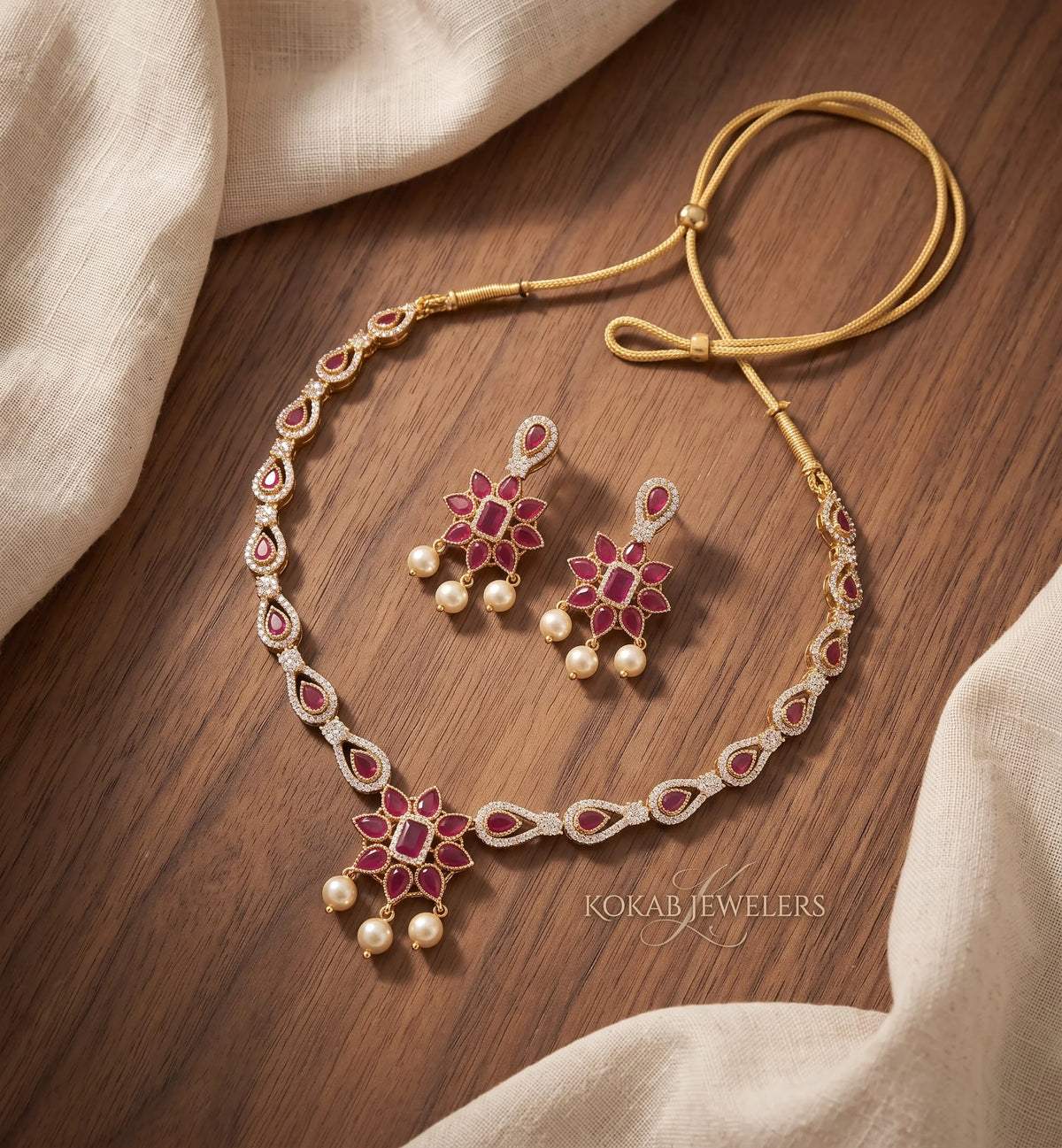 Gold-toned traditional necklace and matching earrings with red floral enameled details and small pearl-like drops, arranged on a wooden surface with draped cream fabric. The necklace has a gold cord and a central pendant, and the earrings match the floral motif.