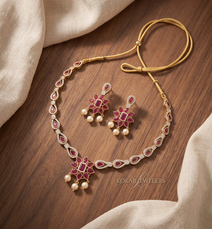 Gold-toned traditional necklace and matching earrings with red floral enameled details and small pearl-like drops, arranged on a wooden surface with draped cream fabric. The necklace has a gold cord and a central pendant, and the earrings match the floral motif.