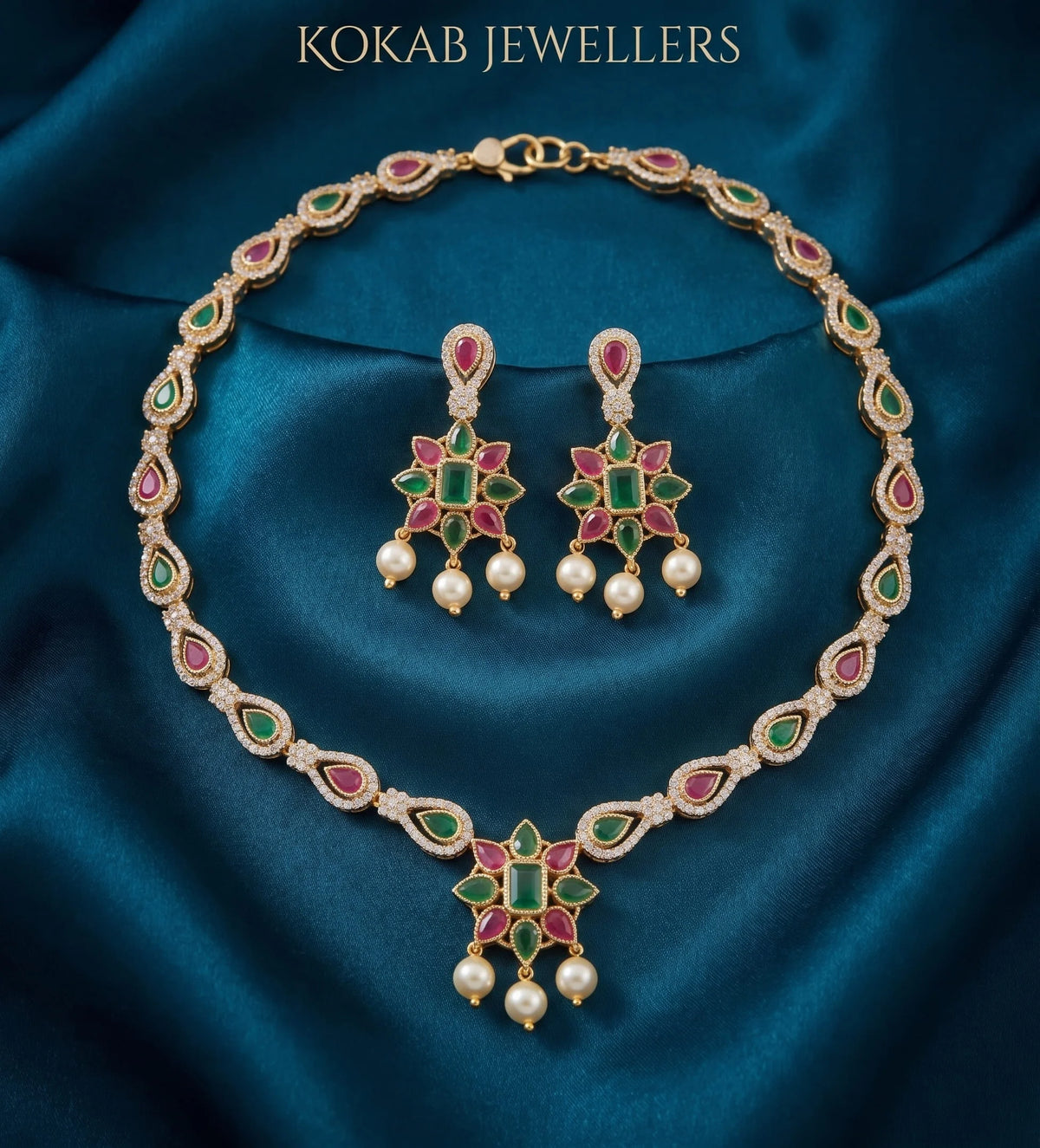 Gold-tone Kokab Jewellers necklace and matching drop earrings with green and pink enamel details, set against a dark teal satin fabric background. The brand name 'KOKAB JEWELLERS' is printed at the top.