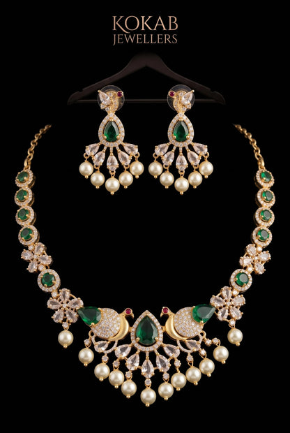 A gold-plated Kokab Jewellers necklace and matching drop earrings set against a solid black background. The jewelry features emerald-cut green stones, round white pearls, and clear pavé-set stones, with the brand name 'KOKAB JEWELLERS' printed in gold at the top.